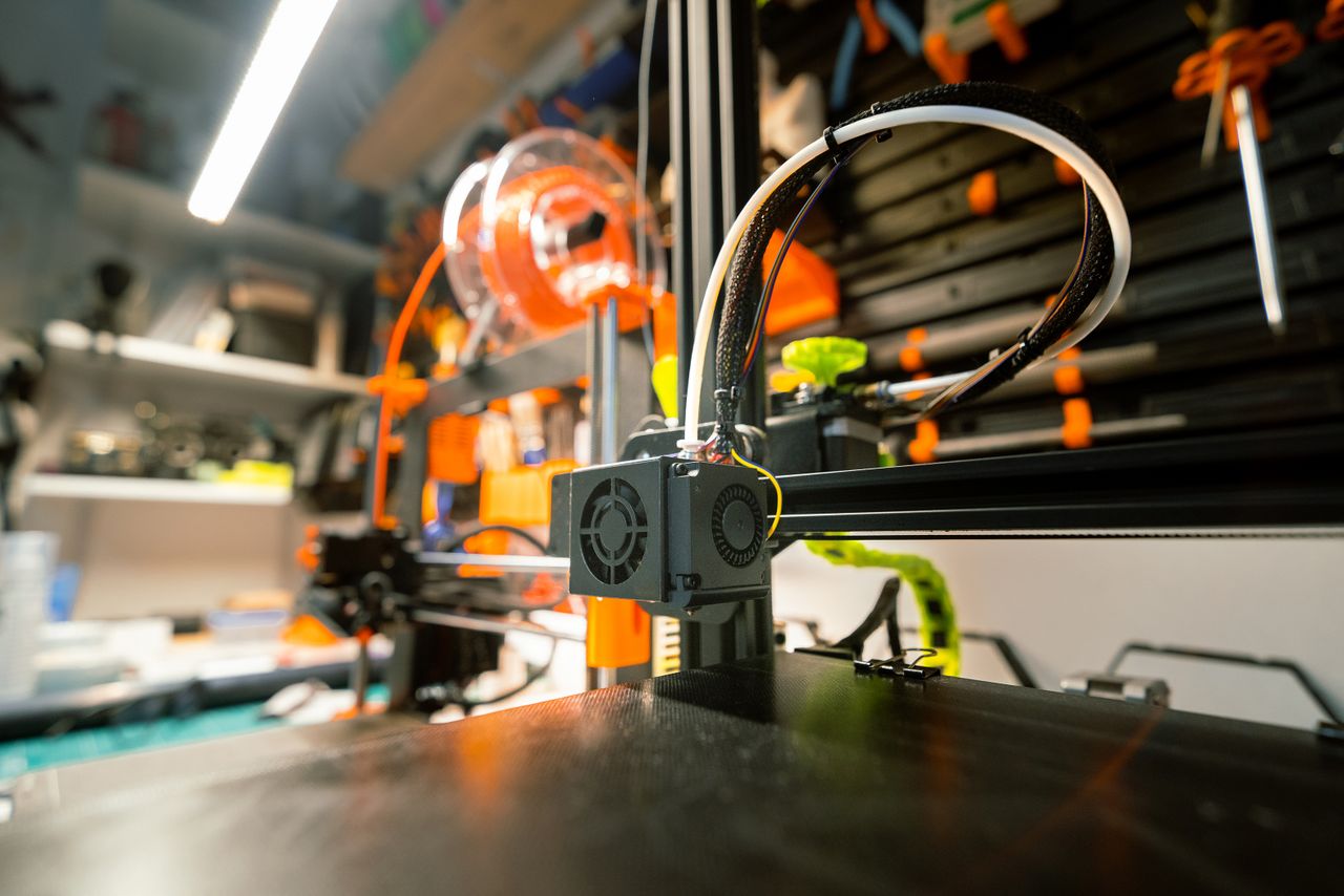 our-story-02 Close-up of a 3D printer in a workshop with visible tools and spool.