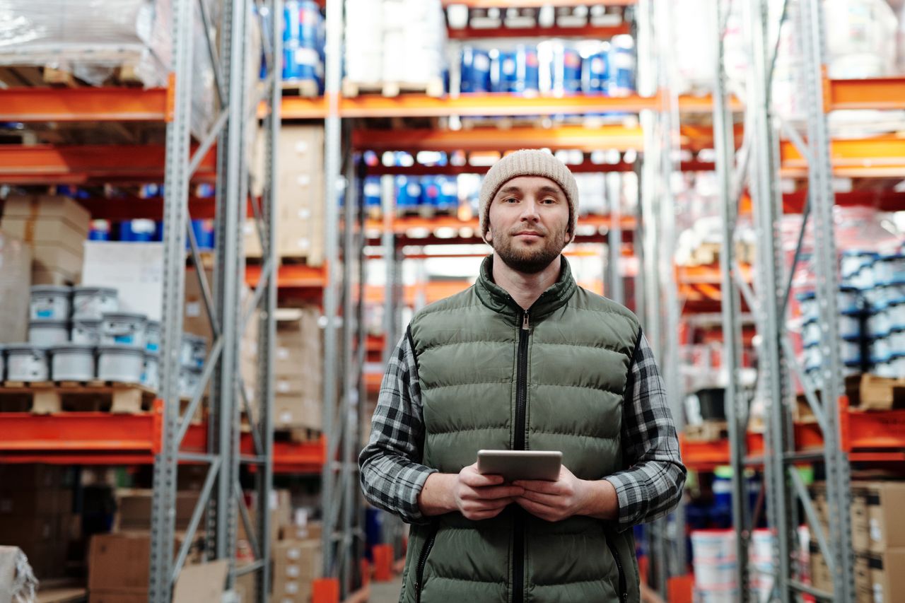 our-mission-01 A warehouse worker wearing a beanie uses a tablet to manage inventory in a storeroom with shelves.